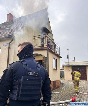 Na zdjęciu umundurowany policjant stoi naprzeciwko budynku mieszkalnego. W tle strażak podczas akcji gaśniczej.