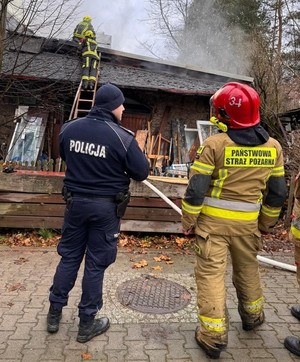 Policjant i strażak przed płonącym budynkiem. Na dachu dwaj strażacy.