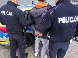 Na zdjęciu dwóch umundurowanych policjantów prowadzi zatrzymanego mężczyznę w kajdankach do radiowozu.