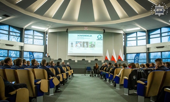 Sala konferencyjna, w niej siedzący na krzesłach mundurowi, pośrodku wyświetlony slajd Konferencja.