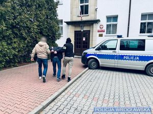 Dwóch funkcjonariuszy prowadzi zatrzymanego mężczyznę w kajdankach do budynku jednostki Policji.