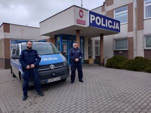 Na zdjęciu dwóch umundurowanych policjantów stoi przy radiowozie. W tle budynek jednostki Policji.