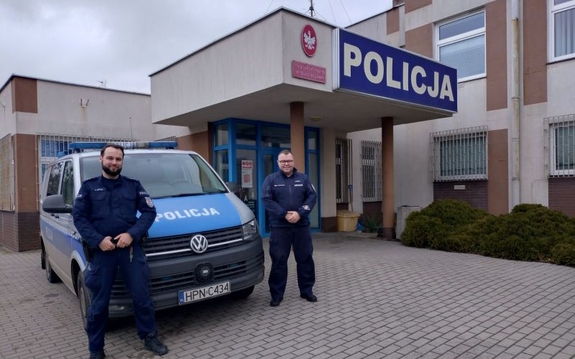 Na zdjęciu dwóch umundurowanych policjantów stoi przy radiowozie. W tle budynek jednostki Policji.