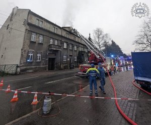 Odgrodzony taśmą teren. W tle wozy strażackie, jeden z nich z wysięgnikiem, na którym znajduje się strażak gaszący budynek.