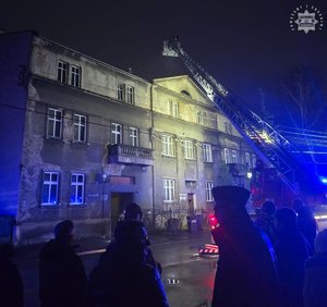 Policjanci i inne osoby stoją przed budynkiem, w którym wybuchł pożar. Dwaj strażacy znajdują się w koszu na wysięgniku wozu strażackiego.