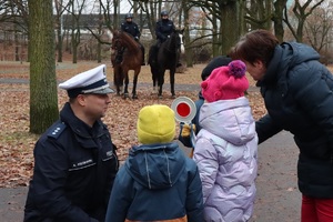 Na pierwszym planie policjant kuca przy trójce małych dzieci i ich opiekunce, w głębi dwaj funkcjonariusze na koniach.