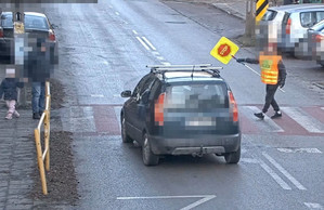 Na zdjęciu samochód osobowy przed przejściem dla pieszych. Na pasach znajduje się mężczyzna ze znakiem STOP. Na chodniku stoi mężczyzna z dzieckiem.