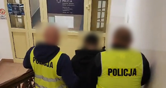 Dwaj nieumundurowani policjanci w żółtych kamizelkach z napisem Policja, prowadzą zatrzymaną osobę.