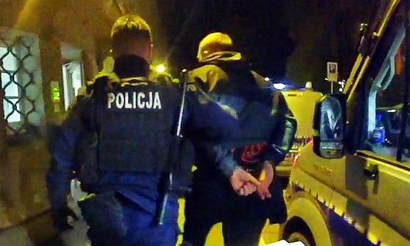 Umundurowany policjant prowadzi zatrzymanego mężczyznę w kajdankach do budynku. Obok stoi zaparkowany radiowóz.