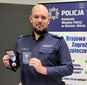 Umundurowany policjant trzyma w ręku odznaczenie i legitymację Honorowego Dawcy Krwi - Zasłużonego dla zdrowia Narodu. Zdjęcie sylwetki od pasa w górę.