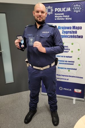 Umundurowany policjant trzyma w ręku odznaczenie i legitymację Honorowego Dawcy Krwi - Zasłużonego dla zdrowia Narodu. Zdjęcie całej sylwetki.