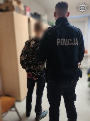 Umundurowany policjant prowadzi zatrzymanego mężczyznę w kajdankach.