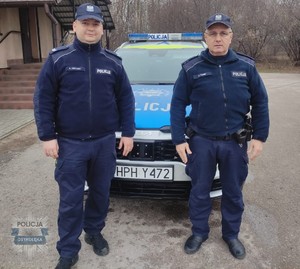 Na zdjęciu dwóch umundurowanych policjantów. W tle radiowóz.