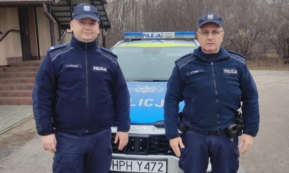 Na zdjęciu dwóch umundurowanych policjantów. W tle radiowóz.