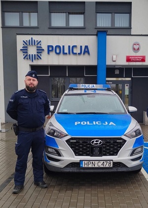 Na zdjęciu umundurowany policjant, w tle zaparkowany radiowóz.