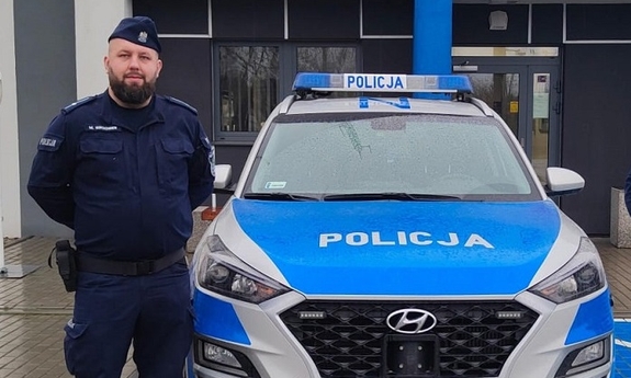 Na zdjęciu umundurowany policjant, w tle zaparkowany radiowóz.