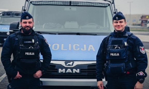 dwóch policjantów stoi przed radiowozem typu bus.