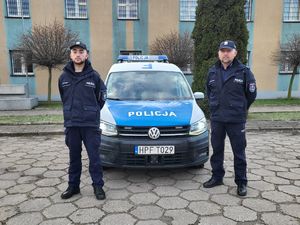 Na zdjęciu dwóch umundurowanych policjantów. W tle zaparkowany radiowóz.