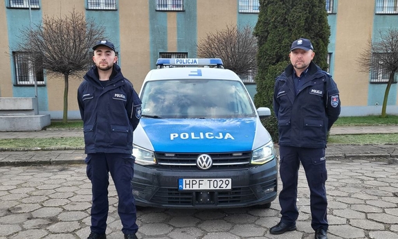 Na zdjęciu dwóch umundurowanych policjantów. W tle zaparkowany radiowóz.