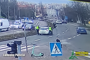 Widok na ulicę. Umundurowani policjanci pomagają kobiecie w białym samochodzie.