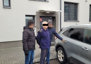 Zatrzymany mężczyzna prowadzony przez dwóch nieumundurowanych policjantów do samochodu.