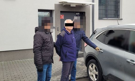 Zatrzymany mężczyzna prowadzony przez dwóch nieumundurowanych policjantów do samochodu.