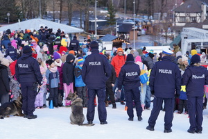 Na zdjęciu umundurowani policjanci z dwoma psami stoją na stoku. Na przeciwko nich znajduje się grupa dzieci z rodzicami. W tle budynki.