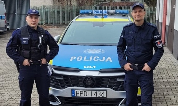 Na zdjęciu dwóch umundurowanych policjantów stoi przy radiowozie.