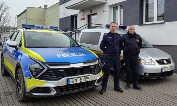 Policjant stoi obok strażnika gminnego, za nimi radiowóz policyjny i pojazd straży gminnej.