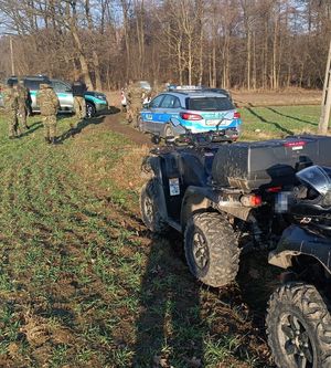 Radiowóz oraz samochód straży granicznej oraz funkcjonariusze na polu w pobliżu terenu leśnego.