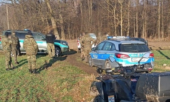 Radiowóz oraz samochód straży granicznej oraz funkcjonariusze na polu w pobliżu terenu leśnego.