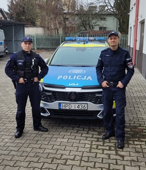 Dwóch policjantów w mundurach stoi po obu stronach radiowozu.