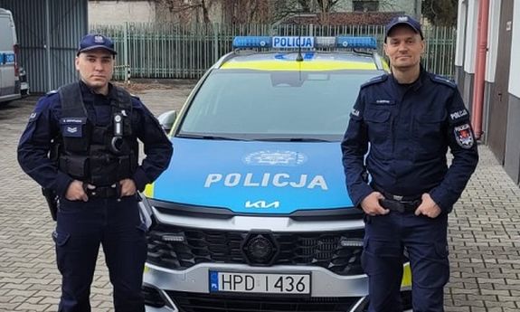 Dwóch policjantów w mundurach stoi po obu stronach radiowozu.