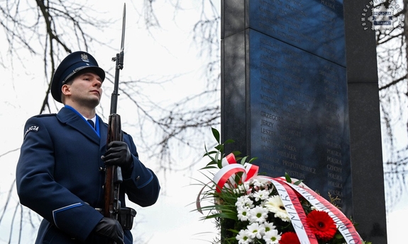 Policjant w mundurze galowym na tle Pomnika upamiętniającego zmarłych w katastrofie hali Międzynarodowych Targów Katowickich.