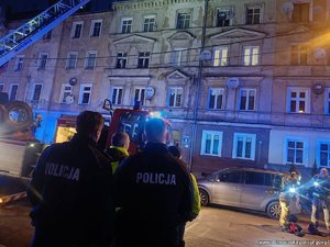 Dwaj policjanci i strażak przyglądają się akcji ratunkowej przy kamienicy.