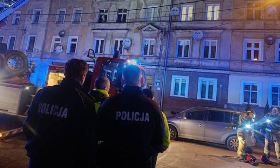 Dwaj policjanci i strażak przyglądają się akcji ratunkowej przy kamienicy.