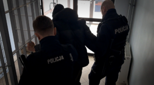 Dwaj umundurowani policjanci prowadzą zatrzymanego mężczyznę.