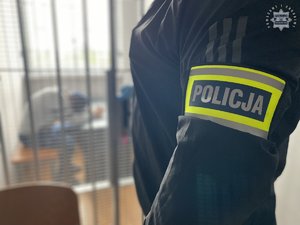 Na zdjęciu część sylwetki policjanta po cywilu z opaską na ramieniu z napisem policja, w oddali w celi zatrzymany.