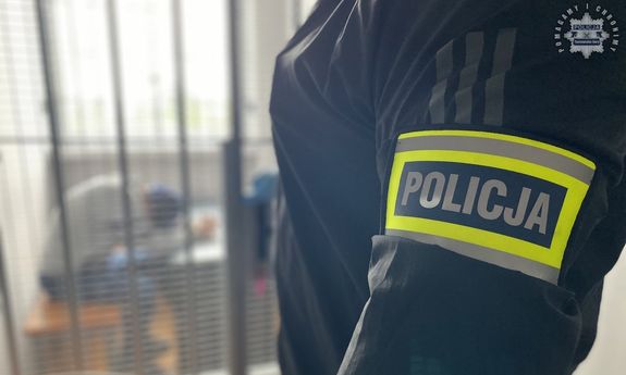 Na zdjęciu część sylwetki policjanta po cywilu z opaską na ramieniu z napisem policja, w oddali w celi zatrzymany.