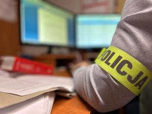 Widoczny łokieć i fragment ramienia policjanta na którym widać opaskę z napisem Policja. W tle widoczne mało wyraźnie komputery.