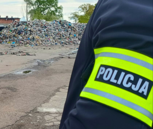 Na pierwszym planie, z prawej strony widoczny fragment ramienia policjanta, na którym znajduje się opaska z napisem Policja. W tle widać wysypisko śmieci.