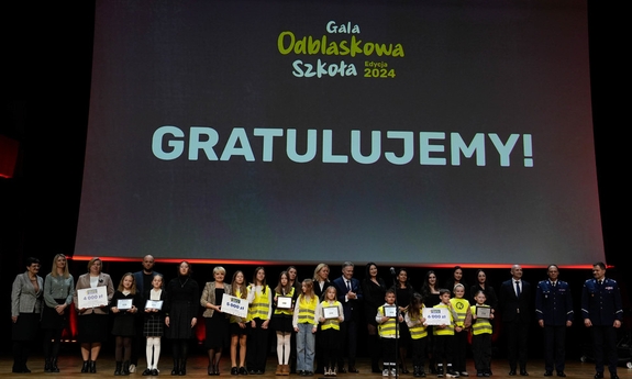 Na tle wyświetlonego baneru gratulujemy stoją uczniowie, nauczyciele z nagrodami wraz z policjantami.