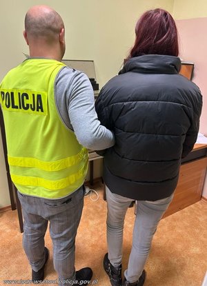 Policjant w żółtej kamizelce z napisem Policja na plecach trzyma za ramię zatrzymaną kobietę.