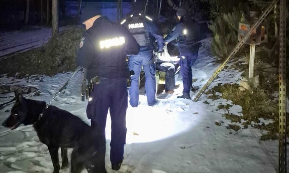 Na zdjęciu umundurowani policjanci i pies policyjny. Dwóch policjantów udziela pomocy odnalezionemu mężczyźnie.