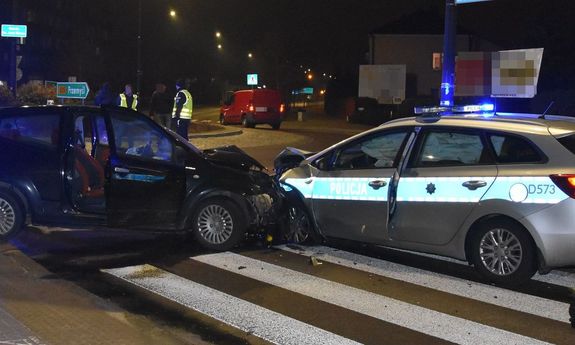 Na drodze uszkodzony radiowóz i samochód osobowy, w tle policjanci i inne osoby.