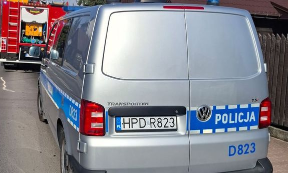 Oznakowany radiowóz policji stojący na ulicy, w tle widoczny wóz strażacki.