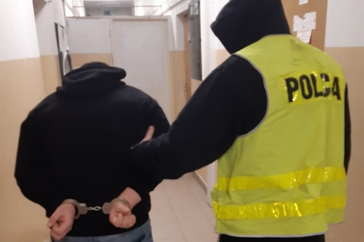 Zatrzymany mężczyzna zakuty w kajdanki prowadzony przez policjanta korytarzem.