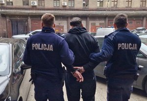 Na zdjęciu dwóch umundurowanych policjantów prowadzi zatrzymanego mężczyznę w kajdankach.