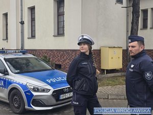 Umundurowani policjant i policjantka stoją przy radiowozie.