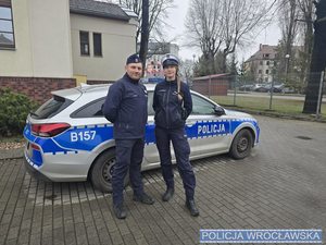 Umundurowani policjant i policjantka stoją przy radiowozie.